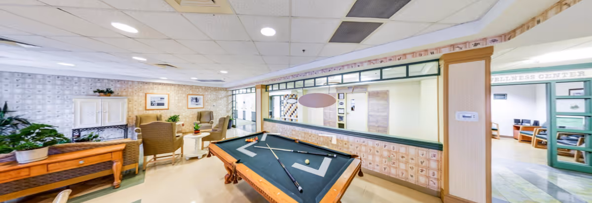Interior view of a recreational room in an assisted living facility featuring a pool table with cues and balls, several armchairs arranged around a small table, potted plants, and a large mirror on the wall. The room has a light-colored floor and ceiling with recessed lighting. A doorway leads to a wellness center visible in the background.