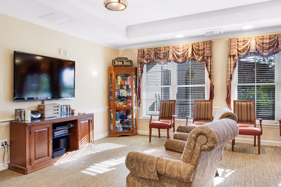 Sunlit senior living common room with armchairs and chairs facing a wall-mounted TV and a small display cabinet.