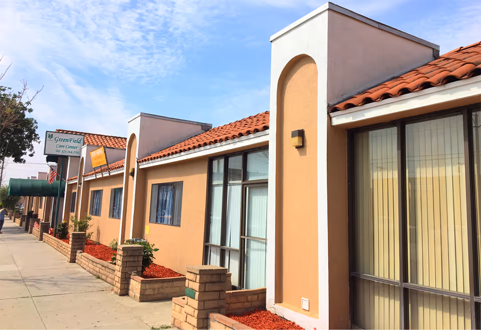 Exterior view of Greenfield Care Center of South Gate, showing a single-story building with beige stucco walls, red tile roof, large windows with vertical blinds, and a sidewalk with small brick planters containing red mulch and some plants.