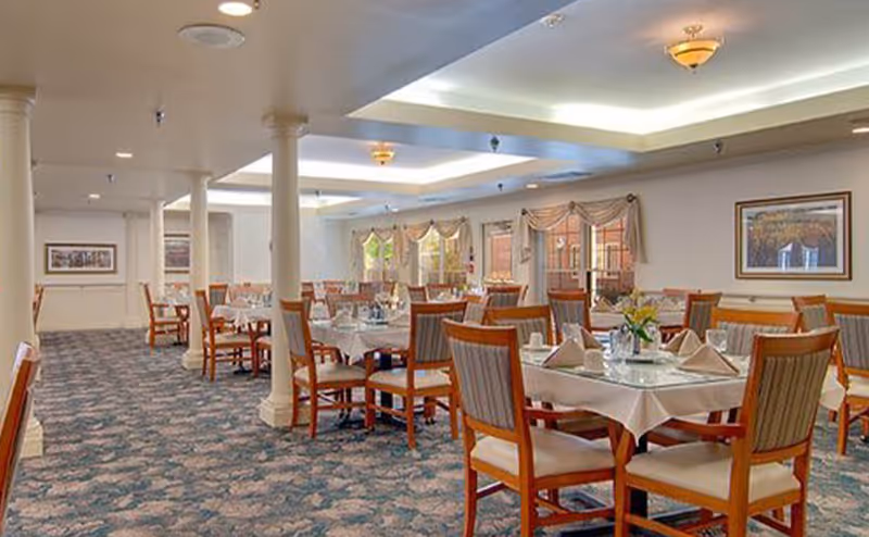 Spacious dining room with multiple tables set for meals, wooden chairs, columns, and patterned carpet.