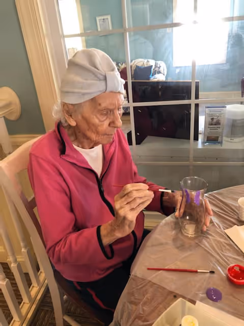 An elderly woman wearing a white head wrap and a pink jacket is sitting at a table painting a glass vase with purple paint. The table is covered with a plastic sheet and has paint supplies including a paintbrush and paint containers. Behind her is a window with a view into another room with a couch and cushions.