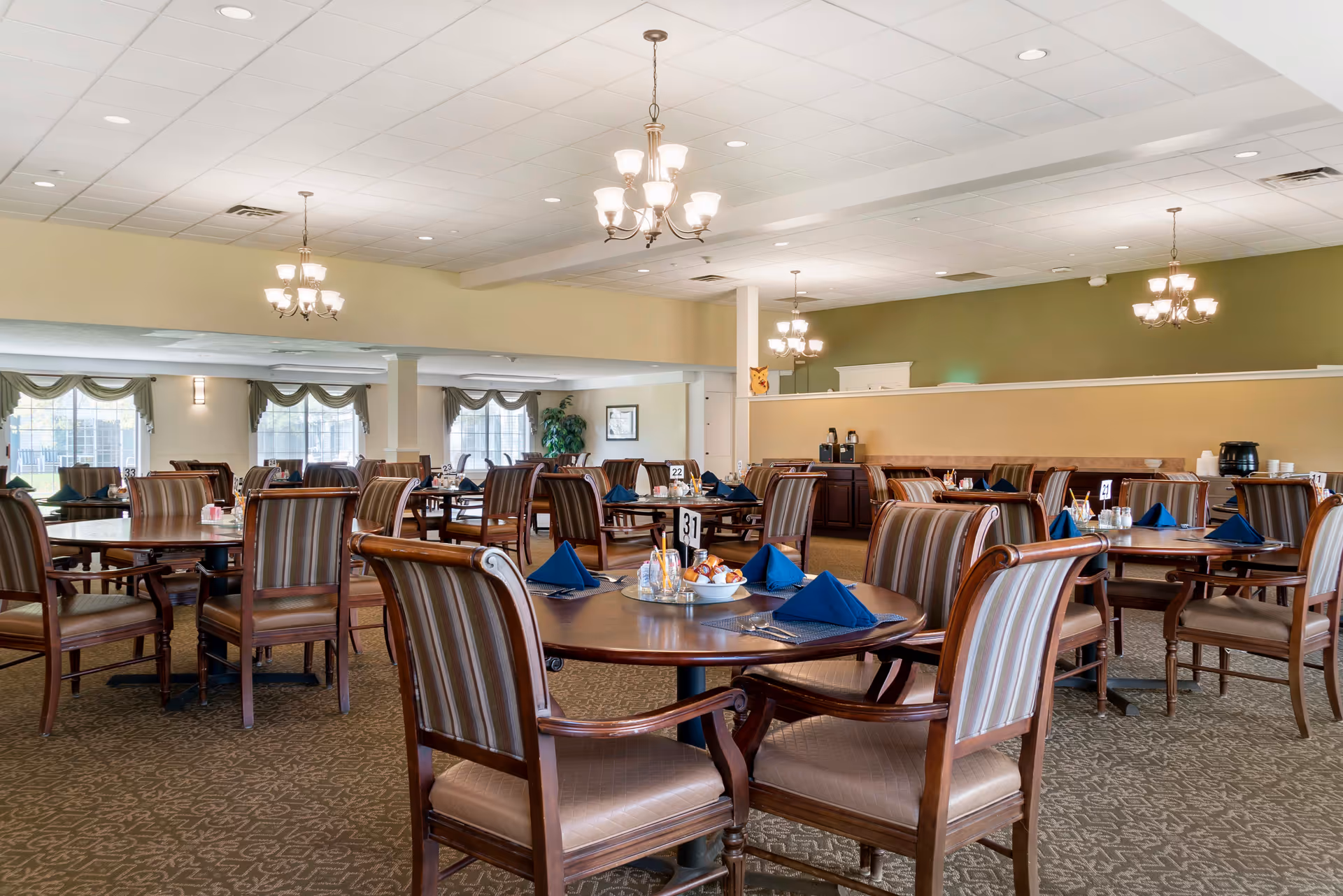 Spacious dining room with multiple round wooden tables and cushioned chairs arranged neatly. Each table is set with blue folded napkins, silverware, and condiments. The room has large windows with green valances, beige walls, and several chandeliers hanging from a white ceiling. There is a buffet area in the background with coffee dispensers and other items.