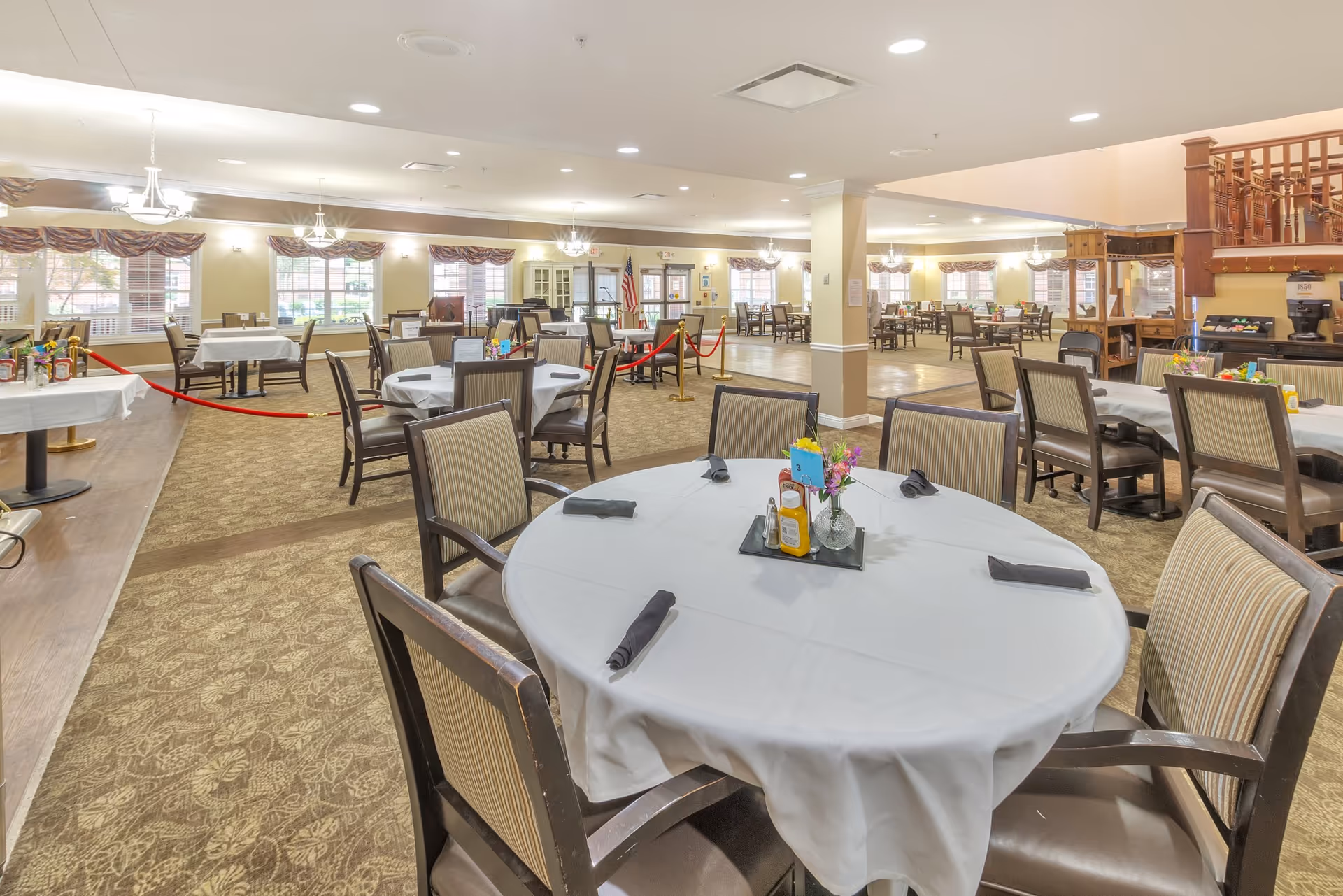 Spacious dining room with round tables set with white tablecloths and chairs in a senior living facility.