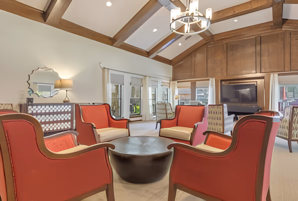 Spacious lounge with a high beamed ceiling, red upholstered armchairs arranged around a round coffee table, large windows and a TV.
