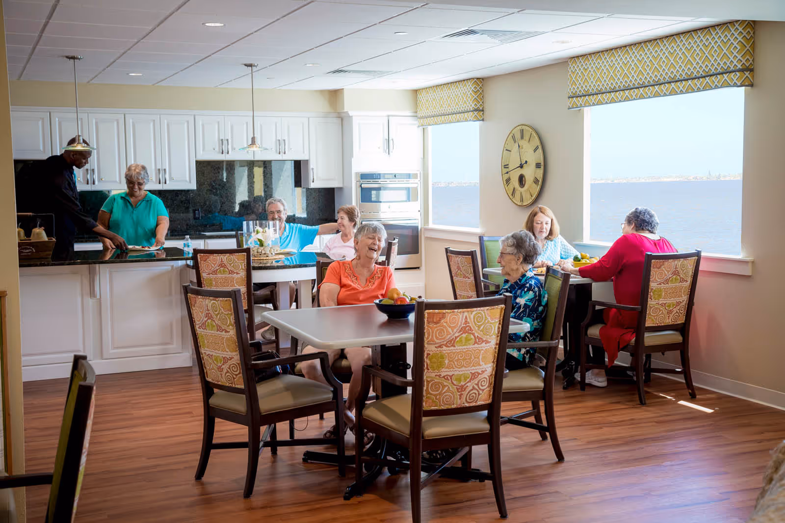 A bright dining area in a senior living facility with several elderly people sitting and chatting around tables. The room features large windows with a view of the water, a clock on the wall, and a kitchen area in the background where a staff member is assisting a resident.