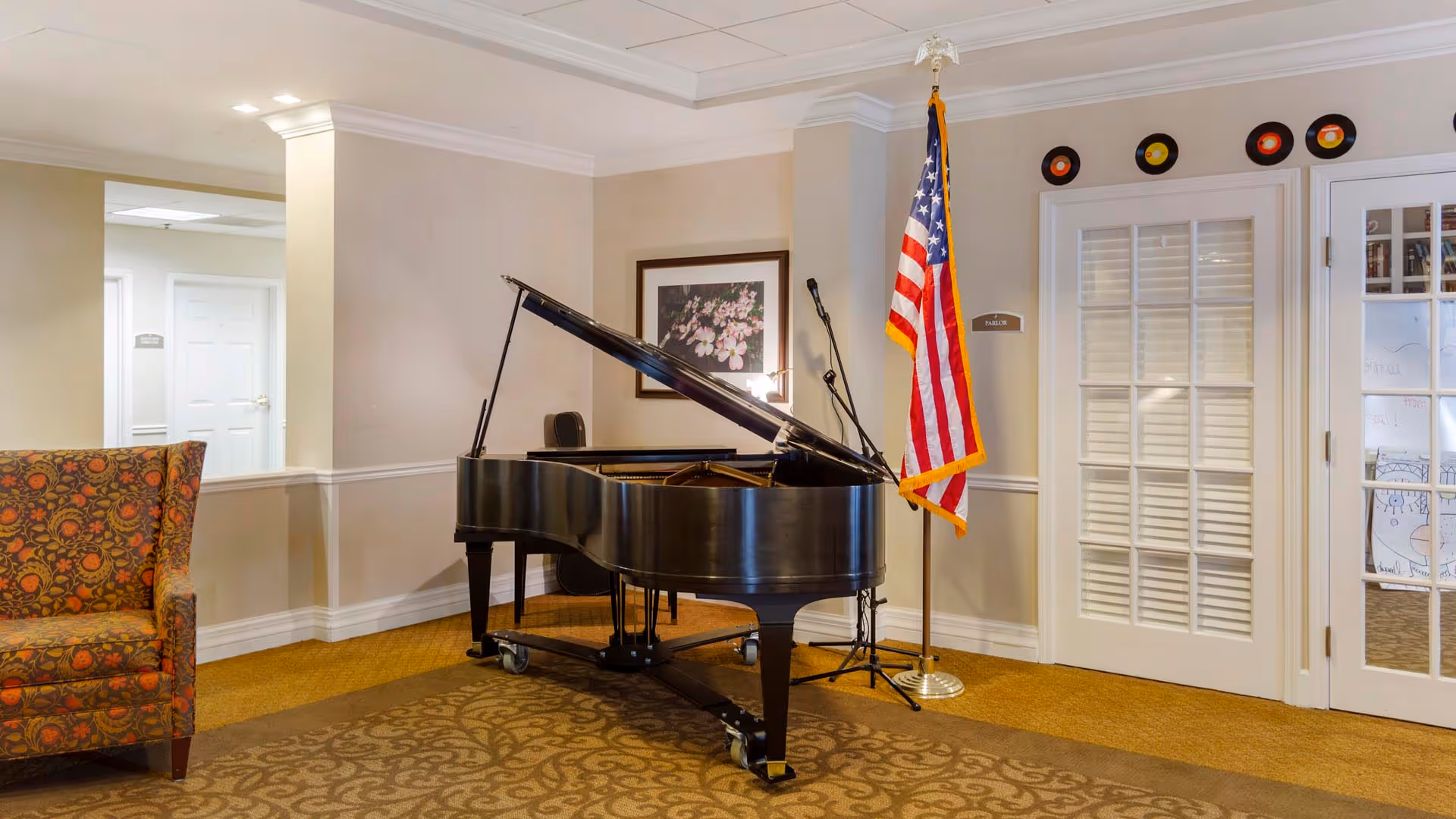 Interior room with a black grand piano on wheels, an American flag on a stand, a floral patterned armchair, a framed picture of flowers on the wall, and a door with glass panels labeled 'Parlor'.