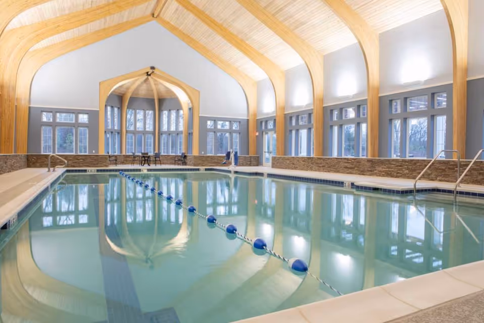 Indoor swimming pool with clear water, surrounded by large windows and wooden arch beams on the ceiling. There are pool safety ropes dividing the pool, and a few tables and chairs are visible near the windows.