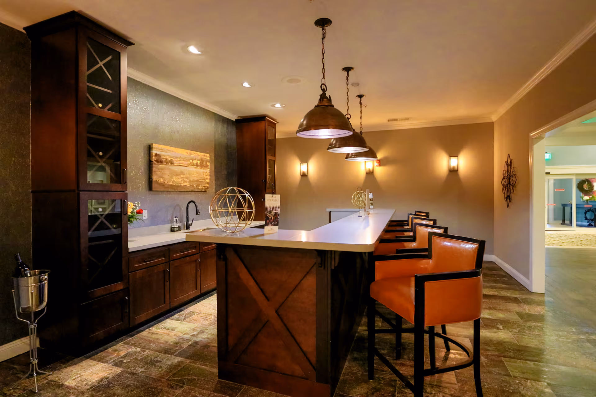 Interior view of a modern kitchen area with a long white countertop island, brown leather bar stools, dark wooden cabinets, pendant lights hanging from the ceiling, and a painting on the wall. The space is warmly lit and has a tiled floor.
