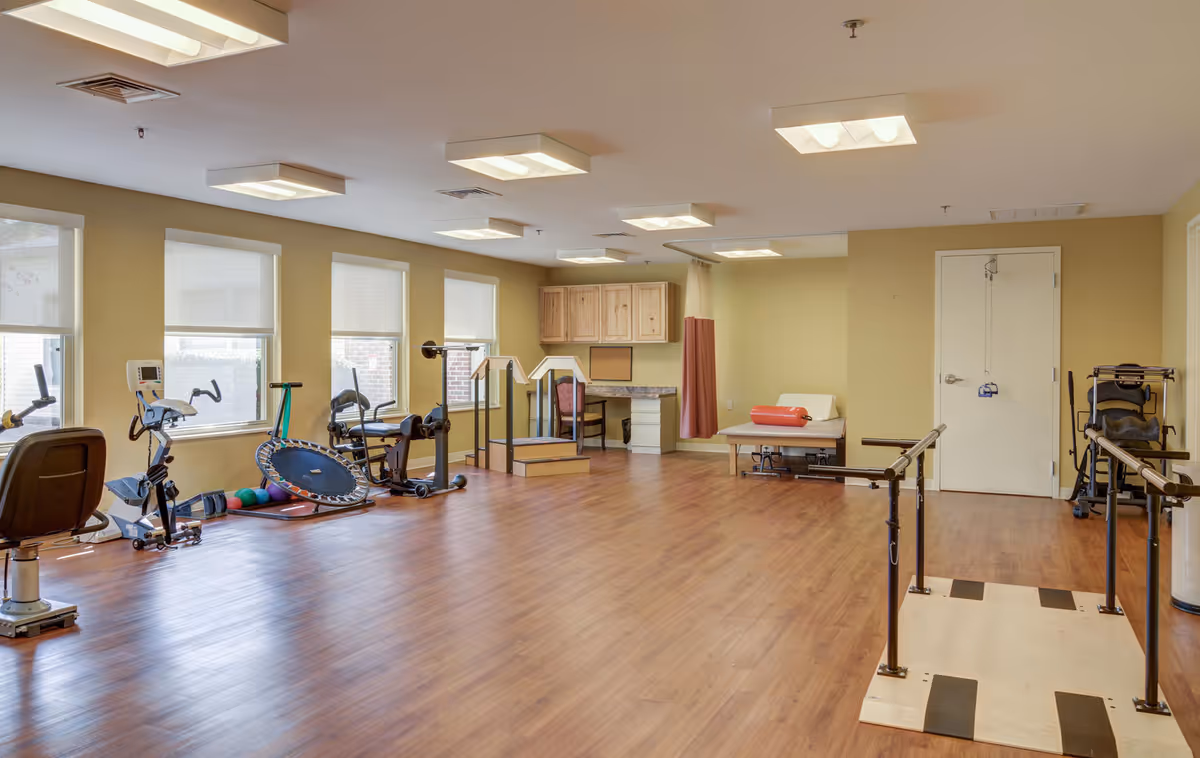 Rehabilitation and fitness room with exercise machines, a small trampoline, parallel bars, a therapy table and windows.