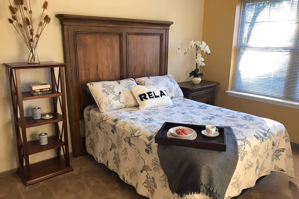 A cozy bedroom with a wooden headboard bed covered in a floral patterned bedspread. There is a white pillow with the word 'RELAX' on it. A wooden tray with a plate of pastries and a teacup sits on a gray blanket at the foot of the bed. To the left, there is a wooden shelving unit with decorative items, and to the right, a wooden nightstand with a white orchid plant. A window with blinds lets in natural light on the right side of the room.