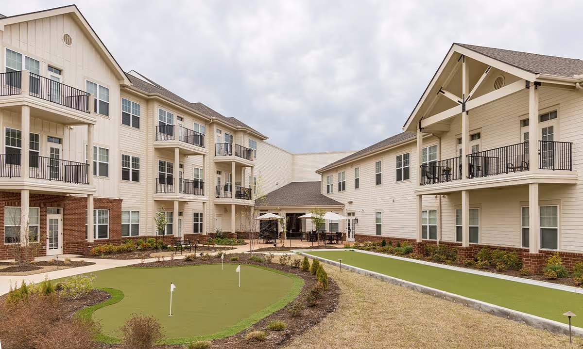 Outdoor courtyard area of a senior living facility with a putting green, surrounded by a two-story building with balconies and patio seating under umbrellas.