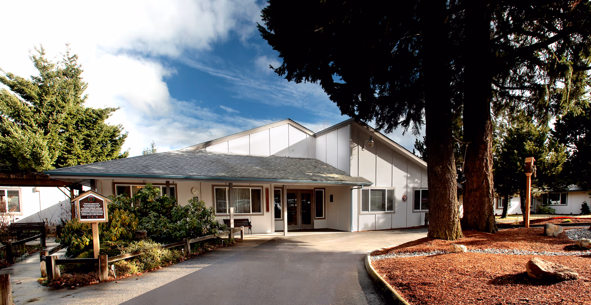 Exterior view of a single-story building with a sloped roof, surrounded by trees and landscaping with mulch and rocks. The building has a covered entrance with glass doors and windows, and a paved driveway leading up to it under a partly cloudy sky.