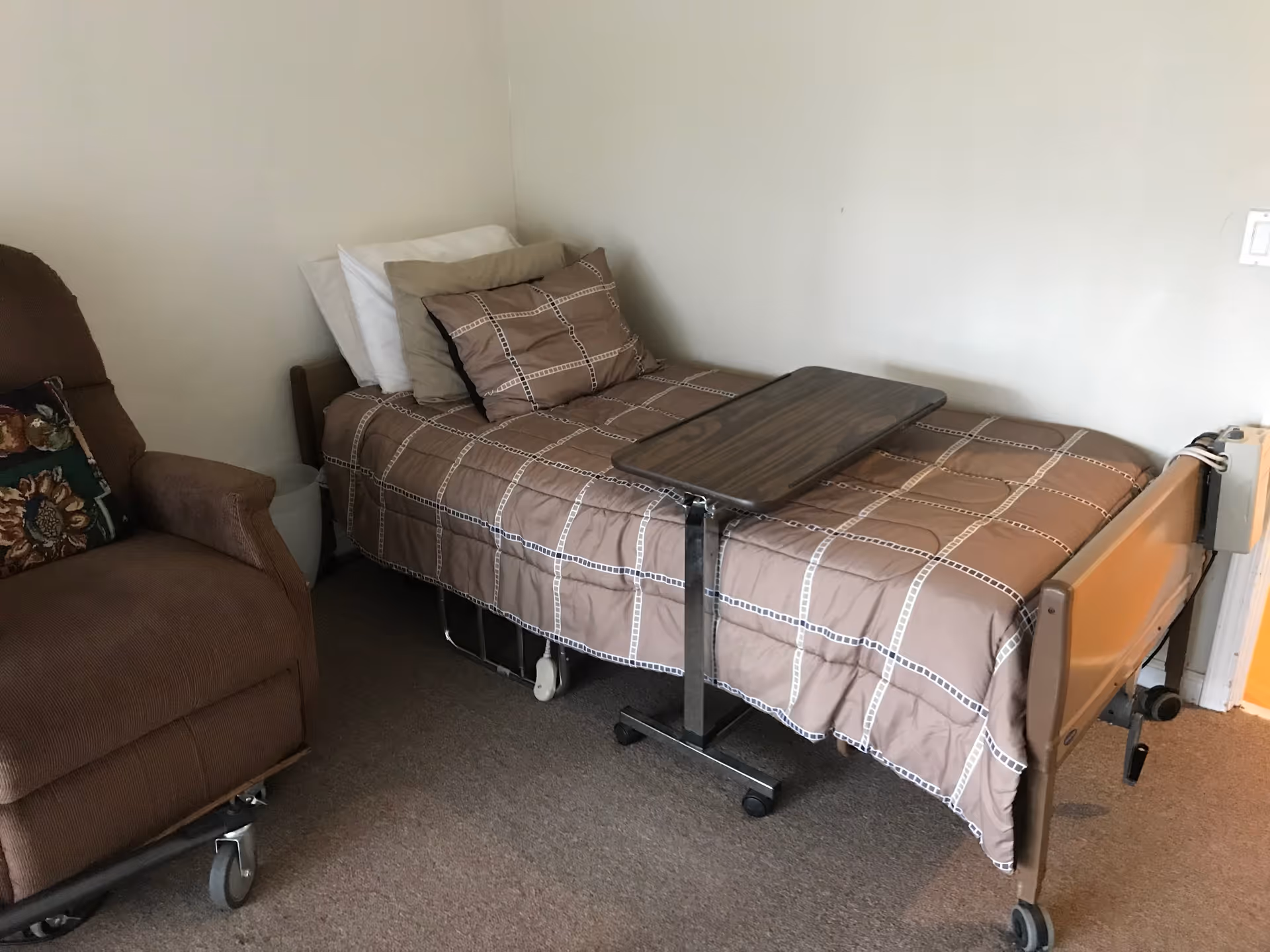 A small room with a single hospital-style bed covered with a brown checkered blanket and multiple pillows. Next to the bed is a brown recliner chair with a floral cushion. A rolling overbed table is positioned over the bed. The walls are plain and light-colored.