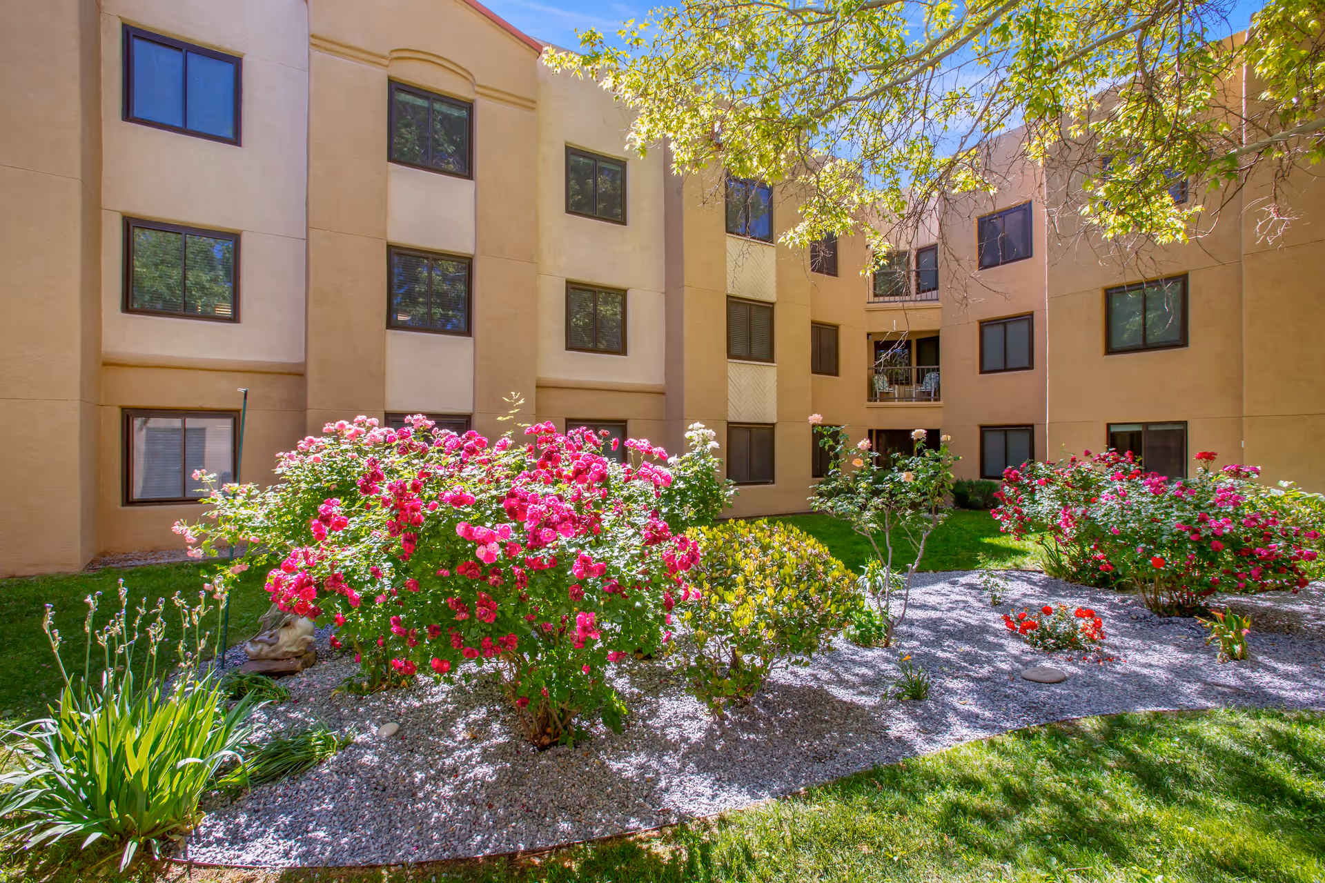 A sunny courtyard garden area with blooming pink and red flowers, green grass, and a few small trees surrounded by a three-story beige building with multiple windows.