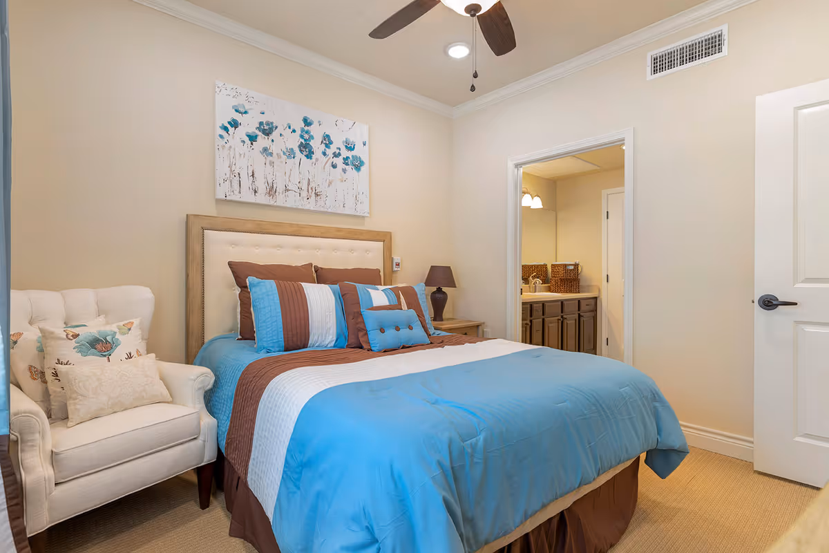 A cozy bedroom with a bed dressed in blue, brown, and white bedding, a beige upholstered headboard, and a white armchair with decorative pillows. A floral painting hangs above the bed. There is a ceiling fan with lights, a nightstand with a lamp, and an open door leading to a bathroom with a sink and mirror.