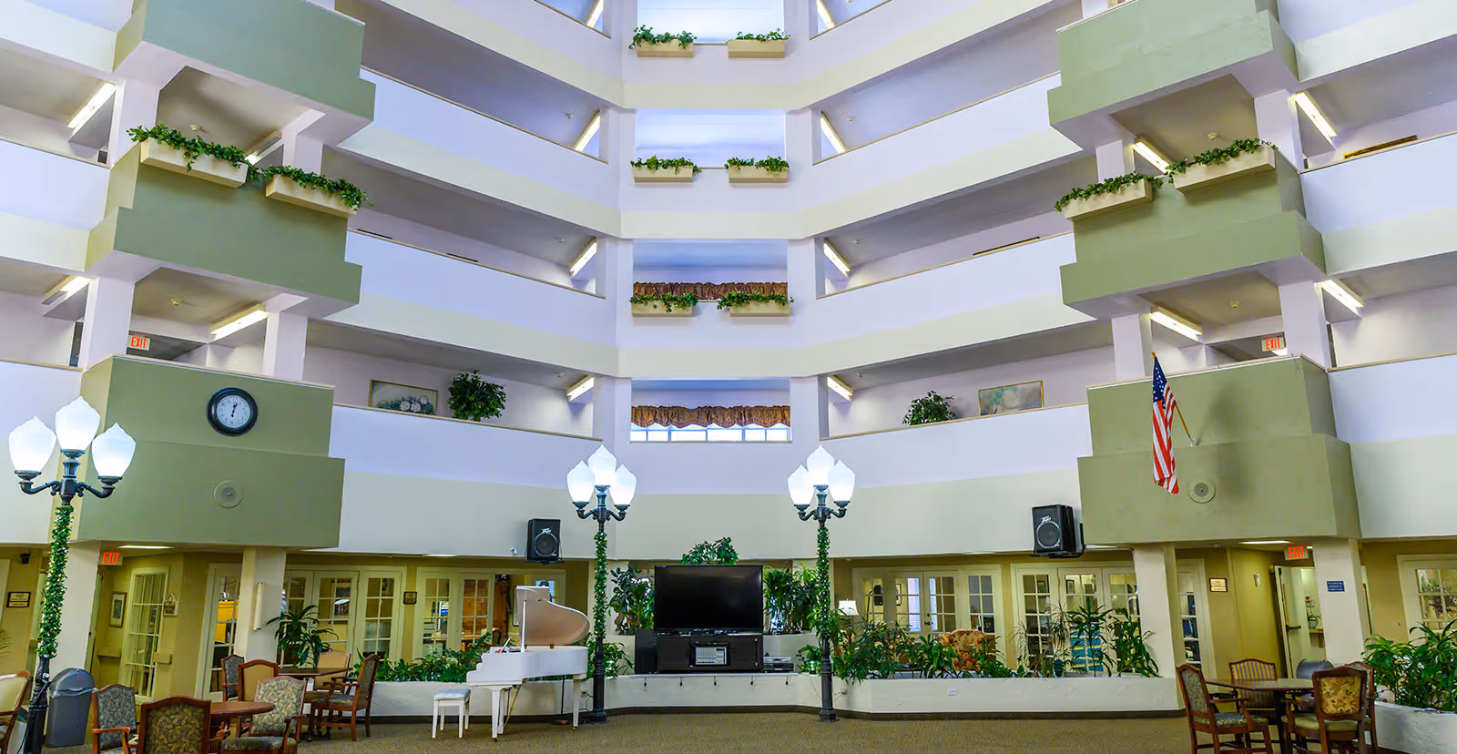Spacious multi-story atrium inside a senior living facility with balconies on each floor decorated with plants. The ground floor features a white grand piano, a large flat-screen TV, several chairs and tables, tall decorative street lamps, and various potted plants. An American flag is displayed on one side of the atrium.