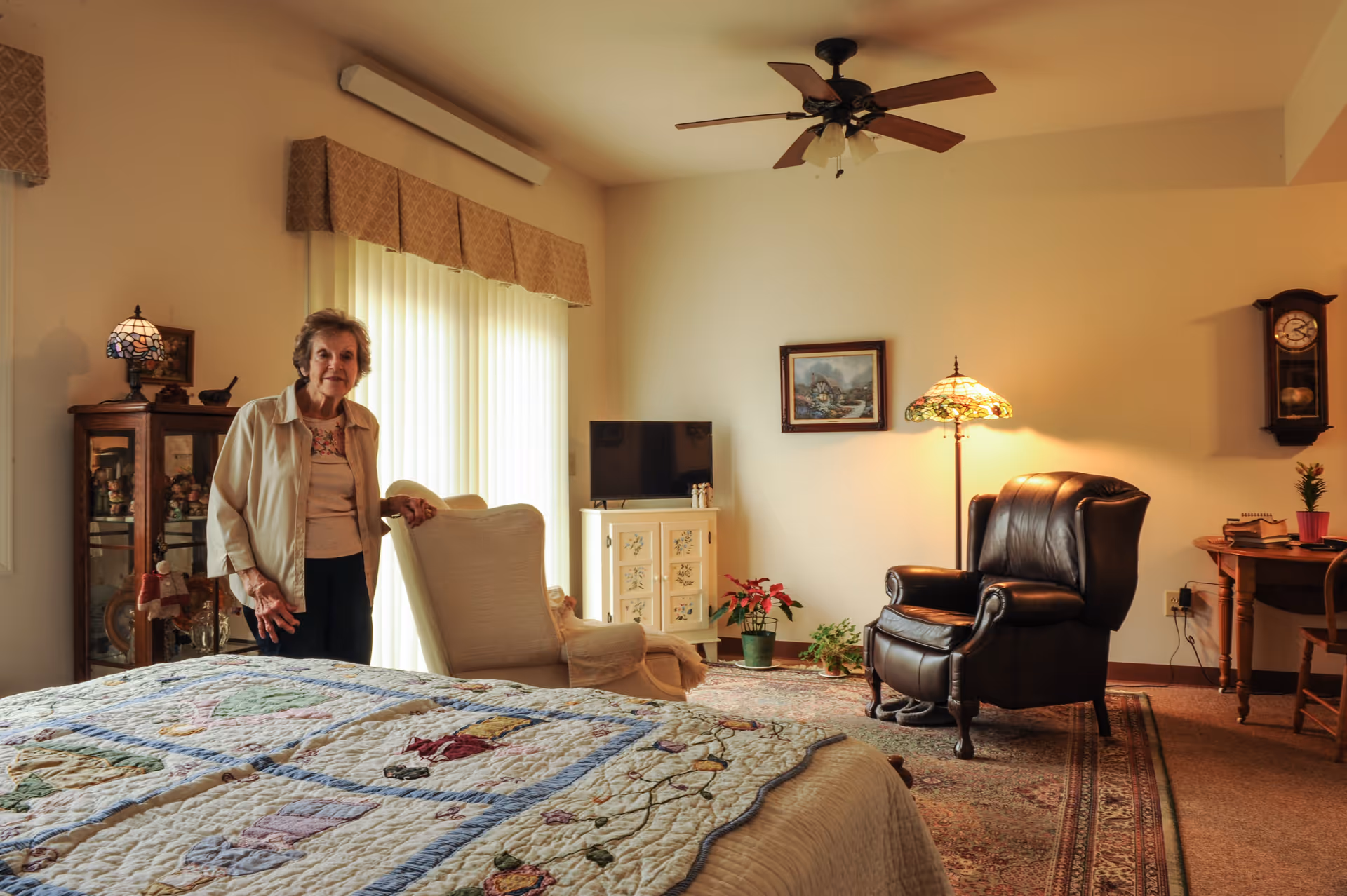 A cozy bedroom in a senior living facility with a quilt-covered bed in the foreground, an elderly woman standing near a cushioned armchair by a large window with vertical blinds, a TV on a small cabinet, a leather armchair, a floor lamp, a wall clock, and a wooden desk with books and a plant.