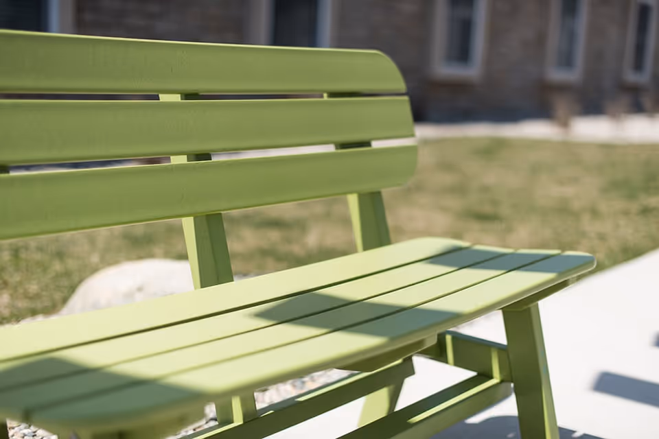 Close-up of a green outdoor bench with a blurred background showing grass and part of a building.