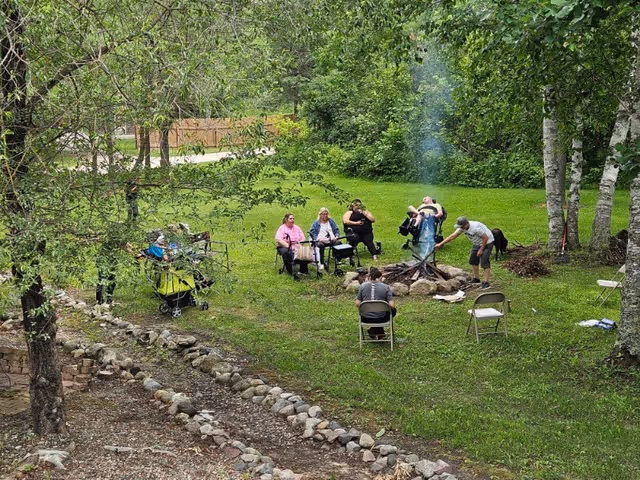 A small group of people sitting on folding chairs around a campfire in a grassy, tree-lined yard.
