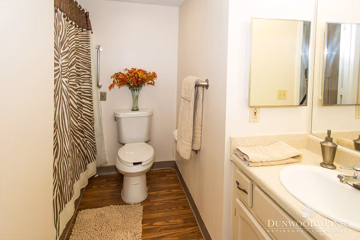 A clean bathroom with a toilet, a shower with a zebra-patterned curtain, a towel hanging on a wall rack, a countertop with a sink, folded towels, and a soap dispenser. There is a vase with orange flowers placed on top of the toilet tank. The floor has a wooden finish and a beige bath mat is placed in front of the toilet.