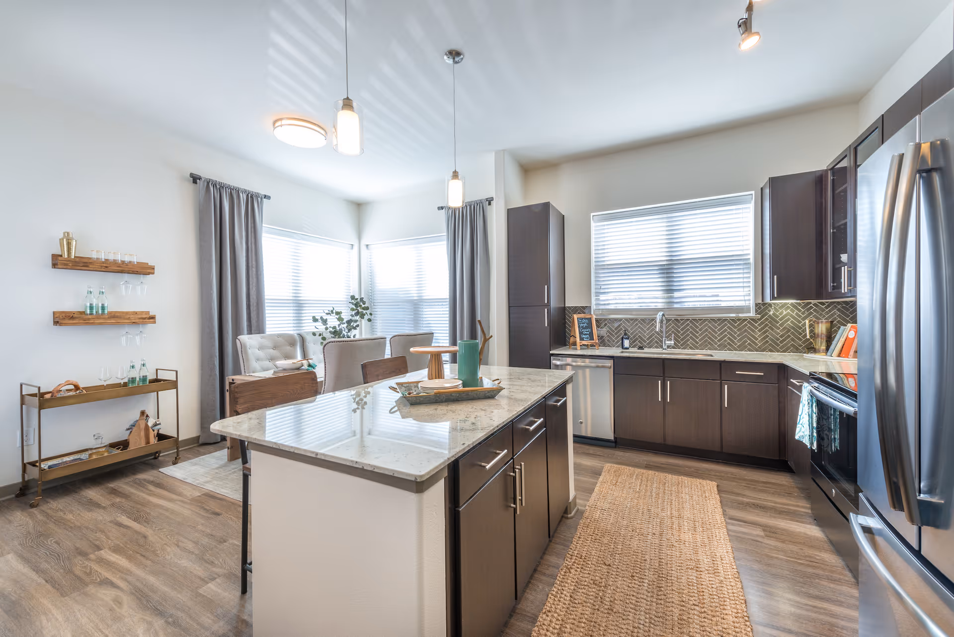 Bright modern kitchen with dark wood cabinets, stainless steel appliances, a large island with a marble countertop, and a dining area with upholstered chairs and a small table near windows with blinds and gray curtains.