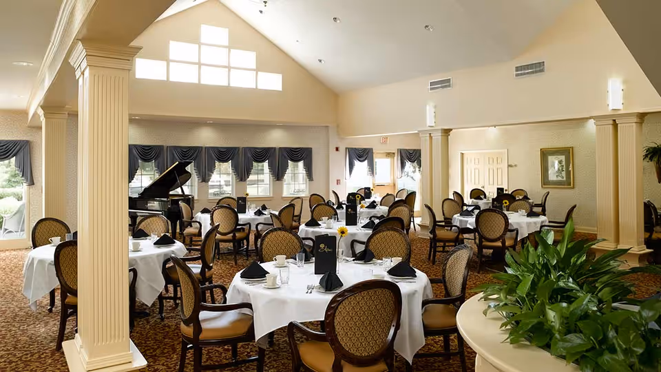 A spacious dining room with round tables covered in white tablecloths, each set with black napkins, cups, and menus. The room features elegant chairs with patterned upholstery, large windows with blue drapes, a grand piano in the corner, and decorative columns. There are plants in the foreground and soft lighting from wall sconces.