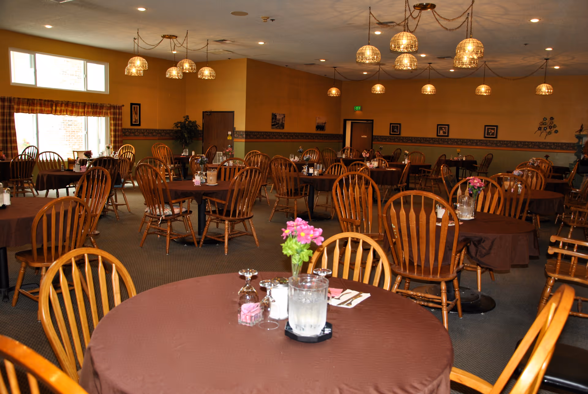 A dining room with multiple round tables covered in brown tablecloths, each set with glasses, napkins, and small flower arrangements. Wooden chairs surround the tables, and the room is warmly lit with hanging pendant lights. Windows with plaid curtains allow natural light to enter the space.