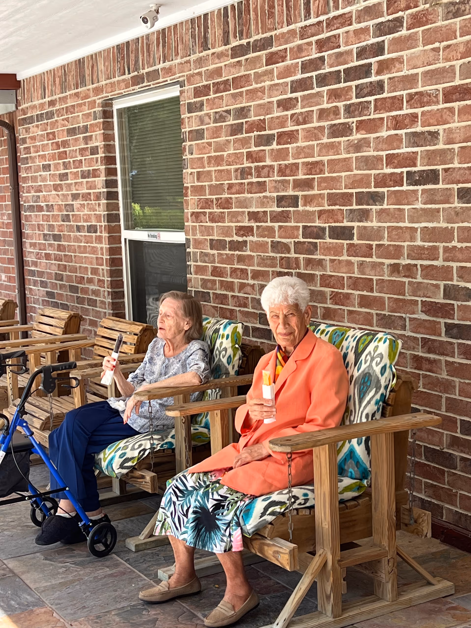 Two elderly women sitting on wooden porch swings with patterned cushions against a brick wall. One woman is holding a bottle of sunscreen and wearing a coral jacket and floral skirt, while the other woman is holding a sunscreen bottle and sitting next to a blue walker.