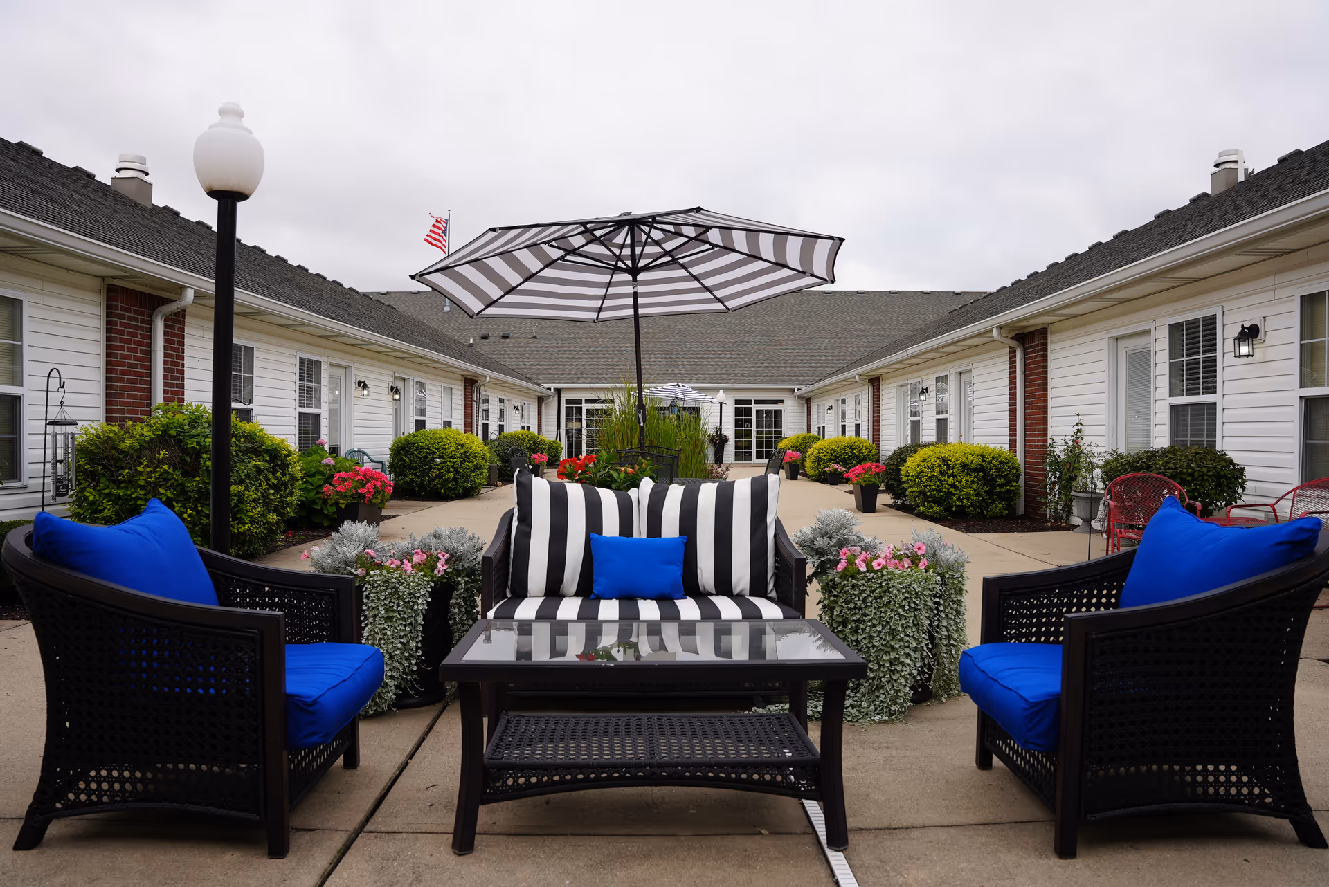 Outdoor courtyard area at Rosegate Commons featuring black wicker patio furniture with blue cushions and a black and white striped umbrella. The courtyard is surrounded by single-story buildings with white siding and brick accents, and decorated with green bushes and colorful flowers.