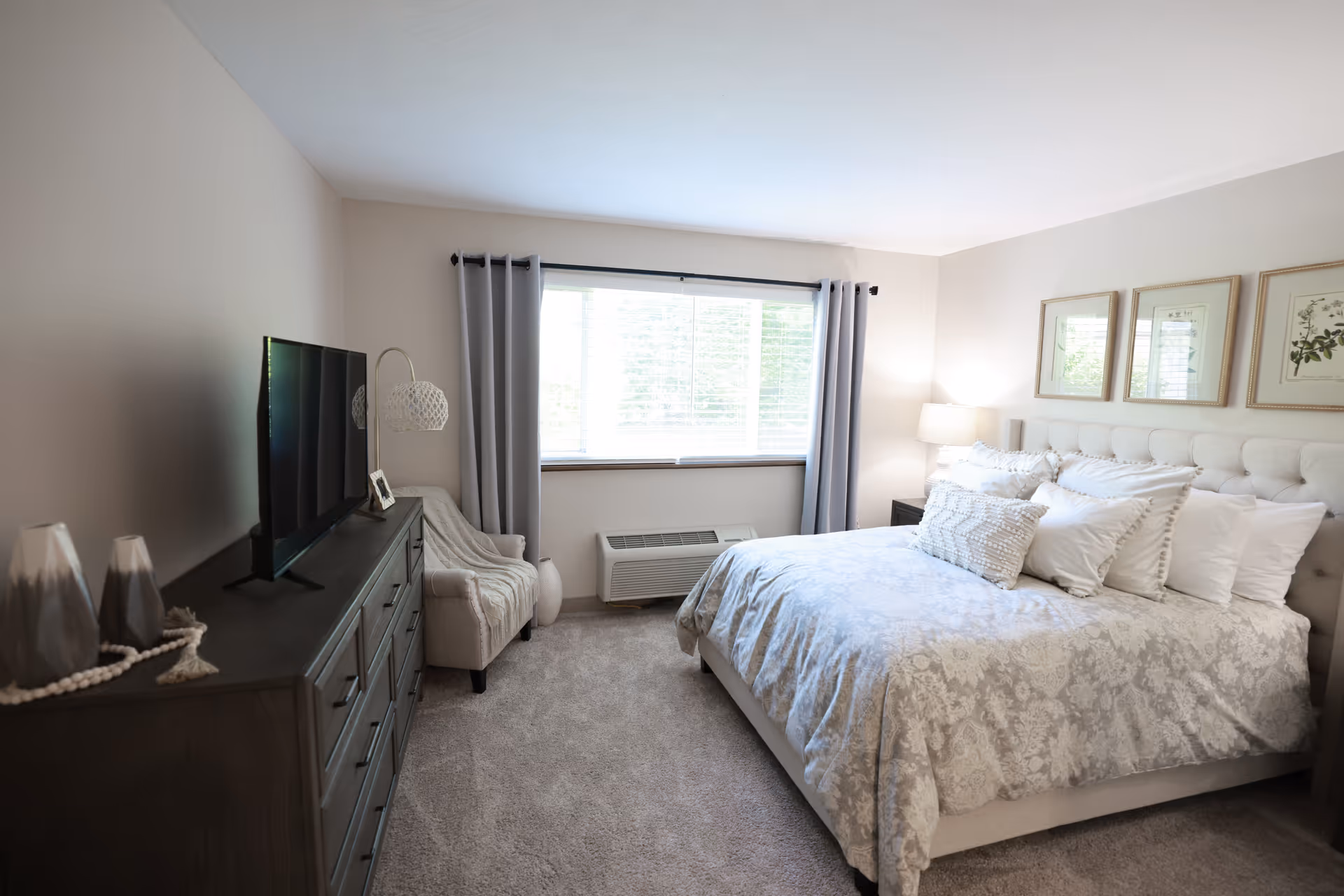 A bright and cozy bedroom with a large bed covered in a light floral-patterned comforter and multiple pillows. There is a window with gray curtains letting in natural light, a cushioned armchair with a throw blanket, a dark wooden dresser with a flat-screen TV on top, and three framed botanical prints hanging on the wall above the bed.