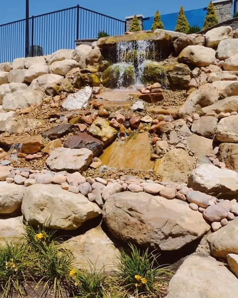 A landscaped outdoor rock waterfall feature with water cascading over large rocks surrounded by smaller stones and some green plants with yellow flowers at the base. A black metal fence and some small evergreen trees are visible at the top of the waterfall.