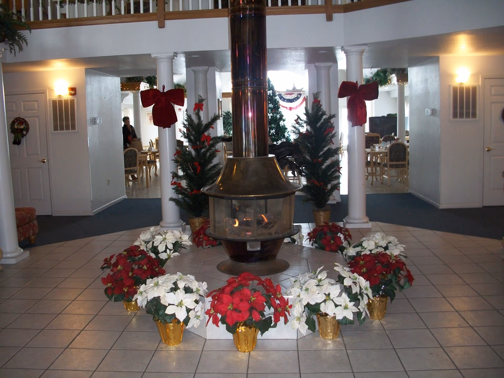 Interior of Cecelia Place Assisted Living decorated for the holidays with a central fireplace surrounded by red and white poinsettias in gold pots. Two small decorated Christmas trees with red bows flank the fireplace. In the background, there are dining tables and chairs, white columns, and holiday decorations including wreaths and garlands.