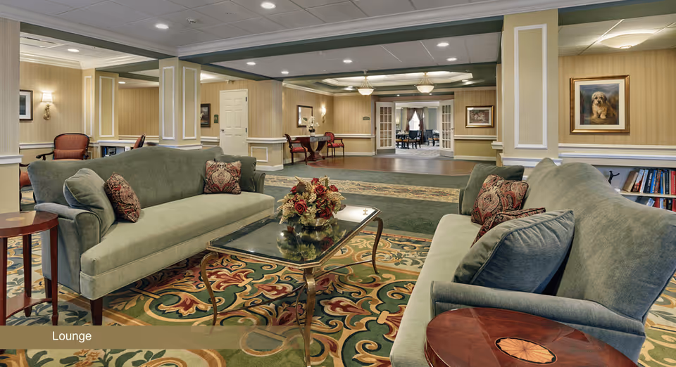 A spacious lounge area with two green upholstered sofas facing each other, a glass coffee table with a floral centerpiece, patterned carpet, and warm beige walls with framed artwork. The room has decorative columns and recessed lighting, with a view into an adjoining room through double doors.