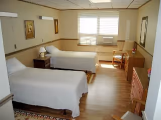 A bright and clean bedroom with two single beds covered in white bedspreads. The room has wooden flooring, a window with blinds letting in natural light, a small nightstand with a lamp between the beds, a wooden dresser, a chair, and framed artwork on the walls.