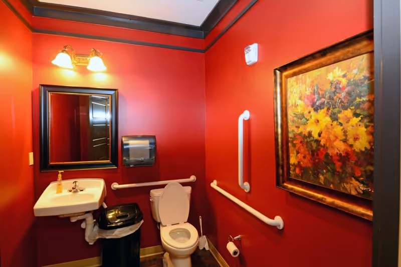 Small red-painted accessible restroom with a toilet, sink, grab bars, mirror, paper towel dispenser and framed floral painting.