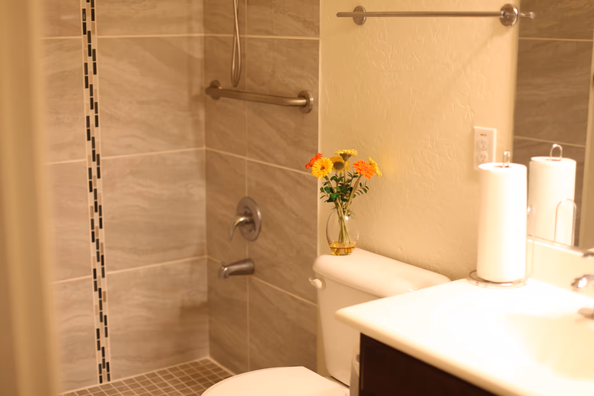 A bathroom with beige tiled shower featuring a grab bar and a handheld showerhead. A white toilet with a small vase of yellow and orange flowers on the tank is next to a sink with a paper towel holder and a mirror above.