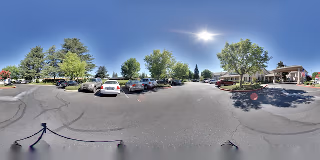 Parking lot and front entrance with parked cars, trees, and a covered porte-cochere under a clear sunny sky.