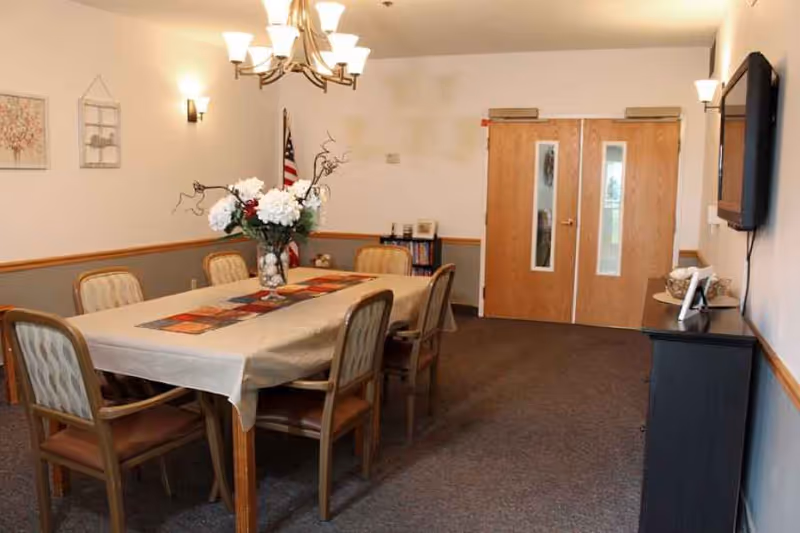 A dining room with a rectangular table covered with a beige tablecloth and a colorful table runner. Six wooden chairs with cushioned seats surround the table. A vase with white flowers and decorative branches is placed in the center of the table. The room has beige walls with a wooden chair rail and gray lower wall paneling. There are two wooden double doors with windows at the far end, an American flag in the corner, framed artwork on the walls, a chandelier overhead, wall sconces, and a flat-screen TV mounted on the right wall above a black cabinet with decorative items.