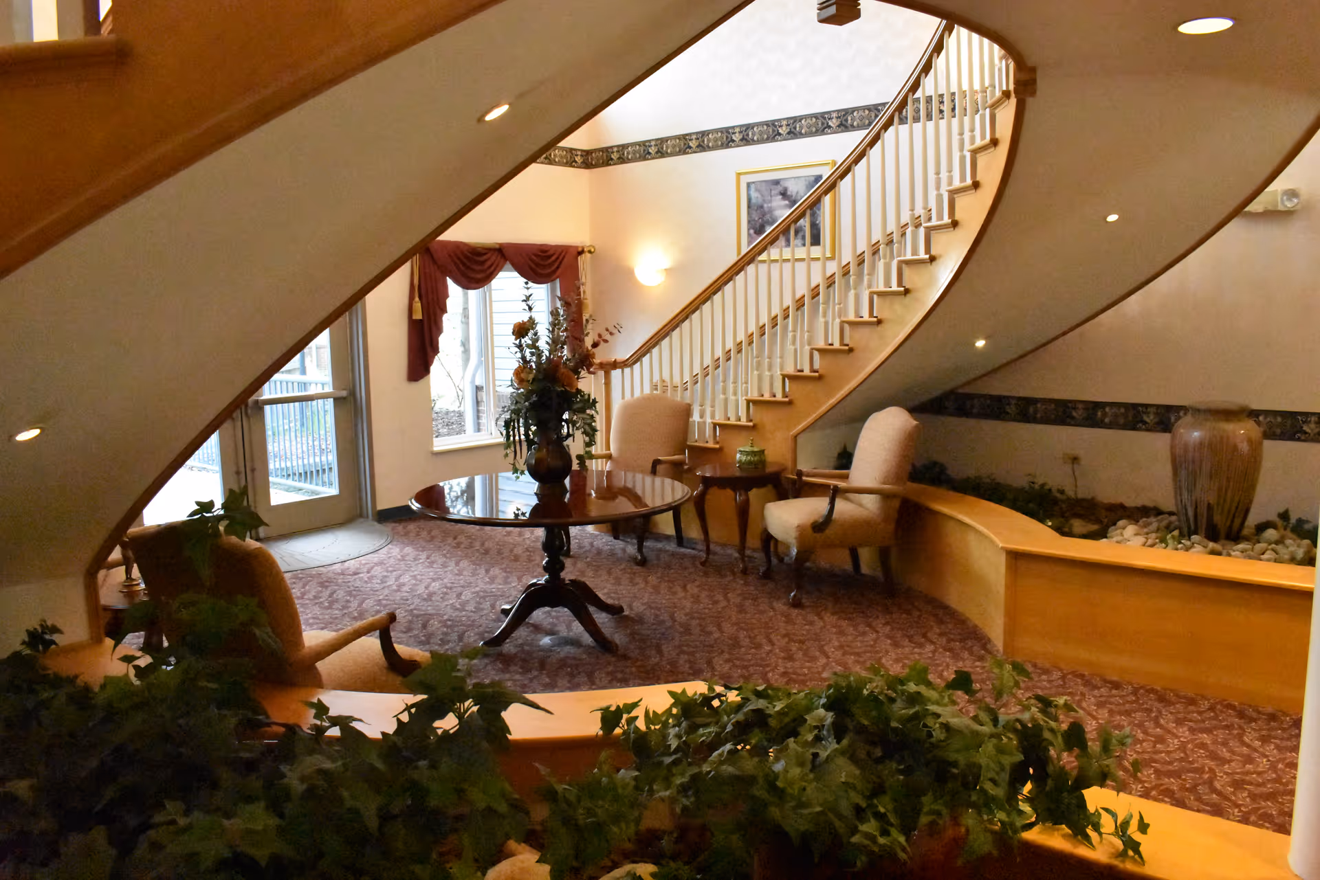 Interior view of a senior living facility lobby area with a curved wooden staircase, two upholstered armchairs, a round wooden table with a floral arrangement, carpeted floor, and decorative plants in the foreground.
