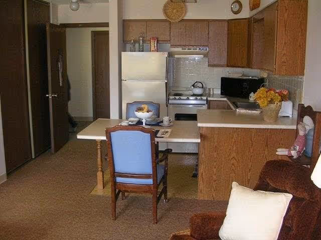 Small kitchen area with wooden cabinets, a white refrigerator, and a stove. A table with two blue cushioned chairs is placed in front of the kitchen counter. A brown armchair with a white pillow is visible in the foreground, and a basket of flowers is on the kitchen counter.