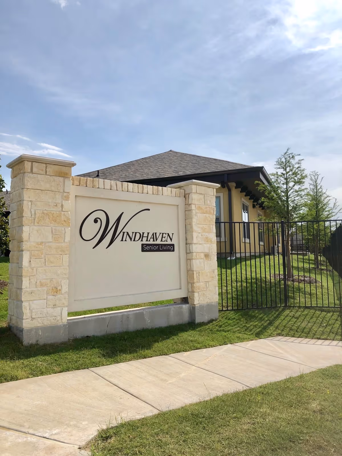 Stone and brick entrance sign for Windhaven Senior Living with a beige building and green lawn behind a black metal fence under a partly cloudy sky.
