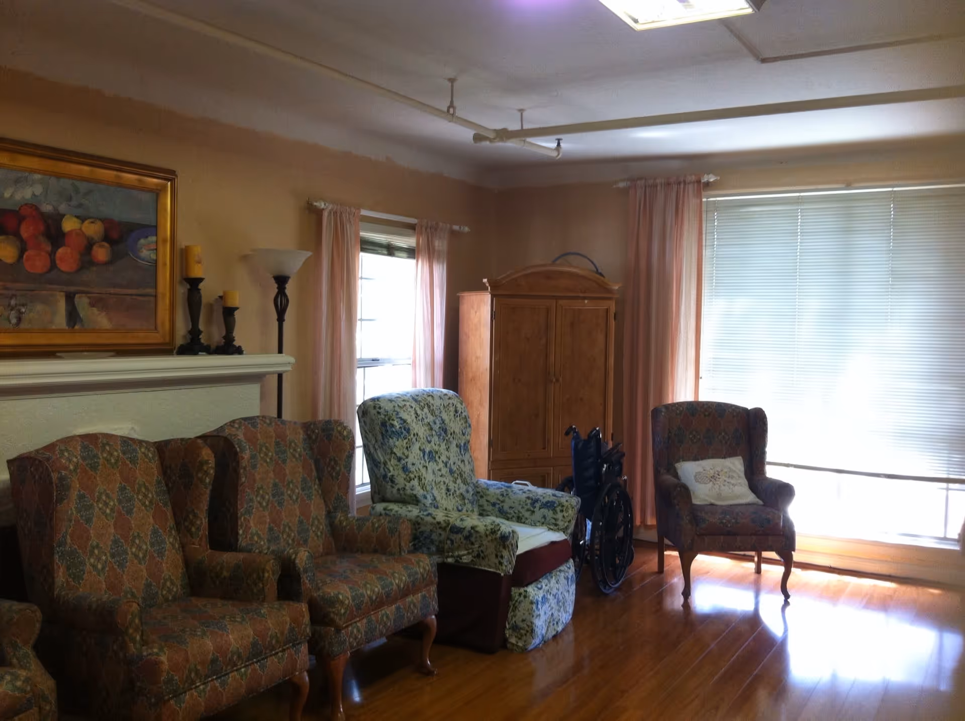 A cozy living room with four armchairs, including two patterned chairs, one floral recliner, and one with a pillow. There is a wooden cabinet in the corner, a wheelchair next to it, two windows with pink curtains, and a large window with blinds letting in natural light. A painting and candles are on the mantel above a fireplace.