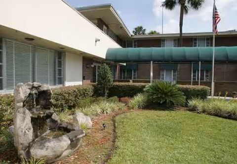 Exterior view of a nursing facility with green awning, landscaped yard, and rock fountain.