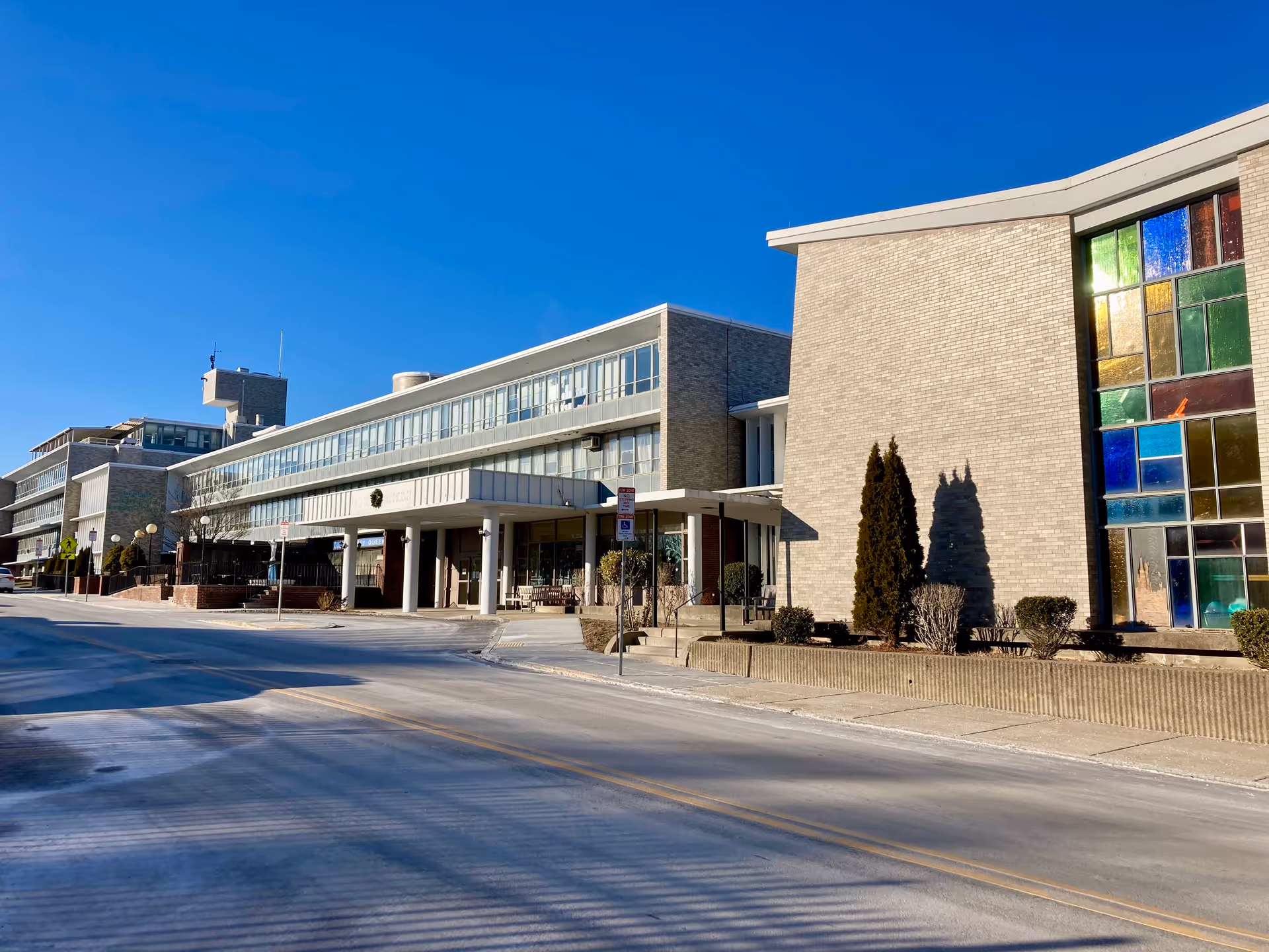 Exterior view of a multi-story building with large windows and a covered entrance, featuring a clear blue sky and a stained glass window on the right side.