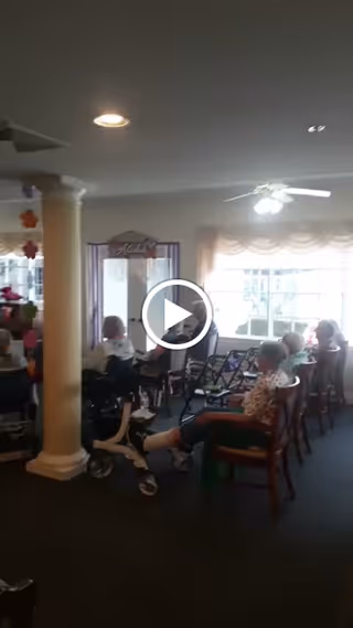 A group of elderly people seated in a common area with chairs and walkers. The room has a ceiling fan, large windows with curtains, and a column near the center. The lighting is soft and natural.