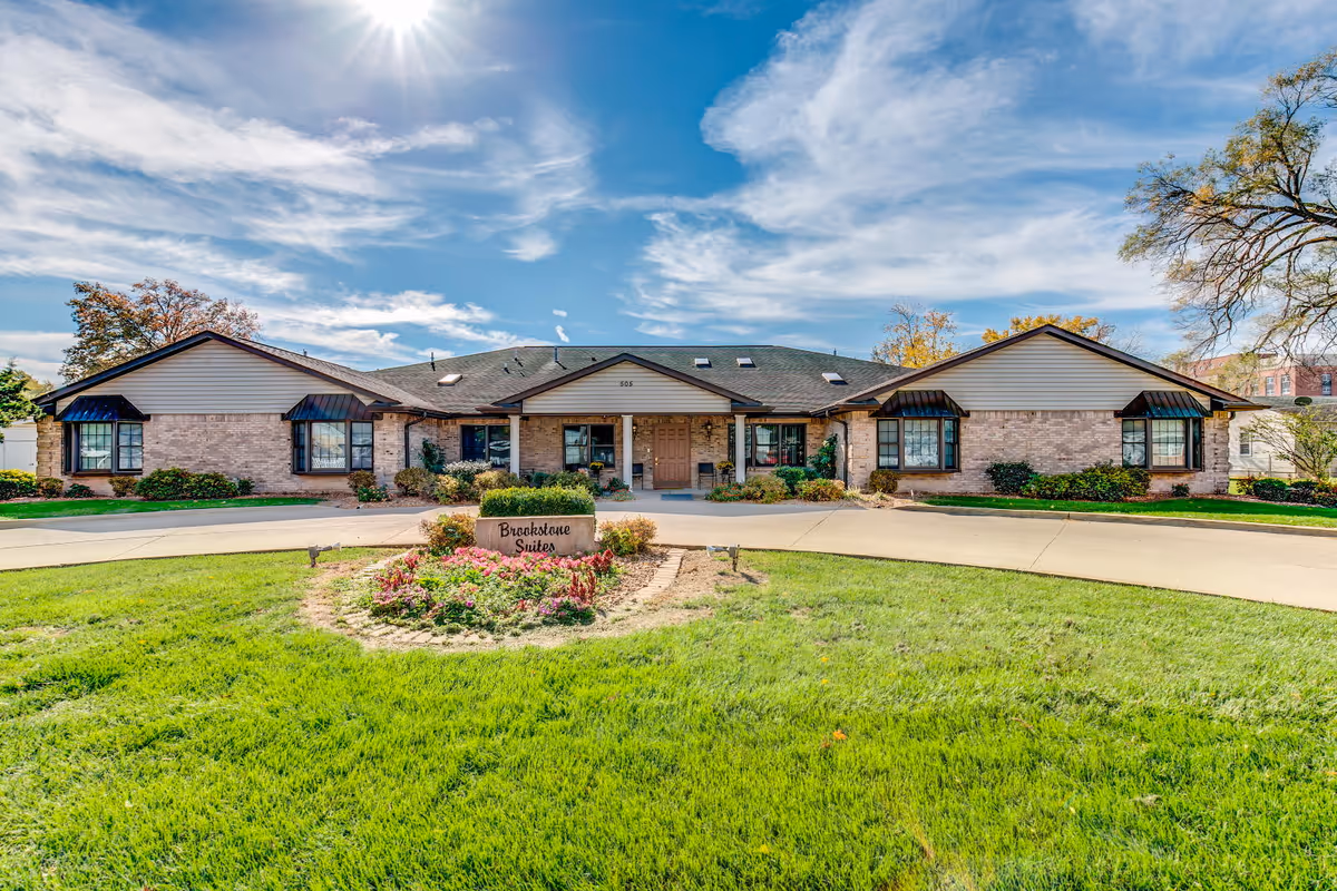 Front exterior view of Brookstone Suites of Effingham, a single-story brick building with a central entrance, surrounded by a well-maintained lawn and flower beds under a partly cloudy blue sky.