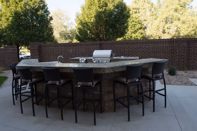 Outdoor patio area with a large stone countertop bar surrounded by eight black bar stools. The bar includes a built-in grill and a small sink. Behind the bar is a brick wall and trees in the background.