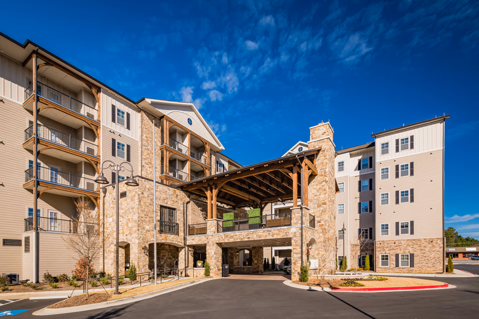 Exterior front entrance of a multi-story senior living building with stone facade, a covered porte-cochère, balconies, and a clear blue sky.