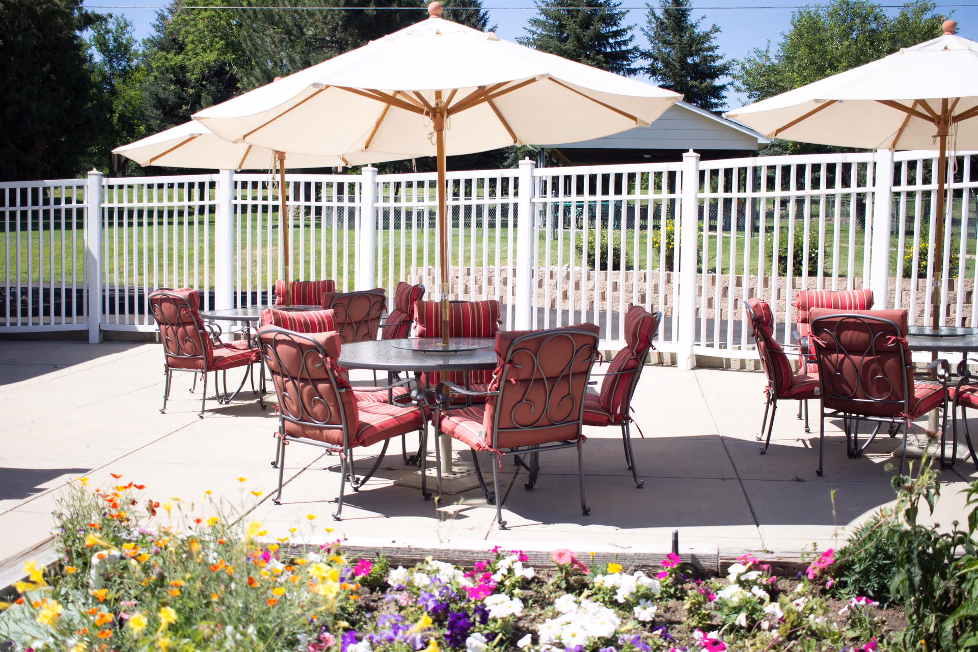 Outdoor patio area with several round tables surrounded by red cushioned metal chairs. Large white umbrellas provide shade over the tables. A white fence encloses the patio, and colorful flowers bloom in the foreground. Trees and greenery are visible in the background.