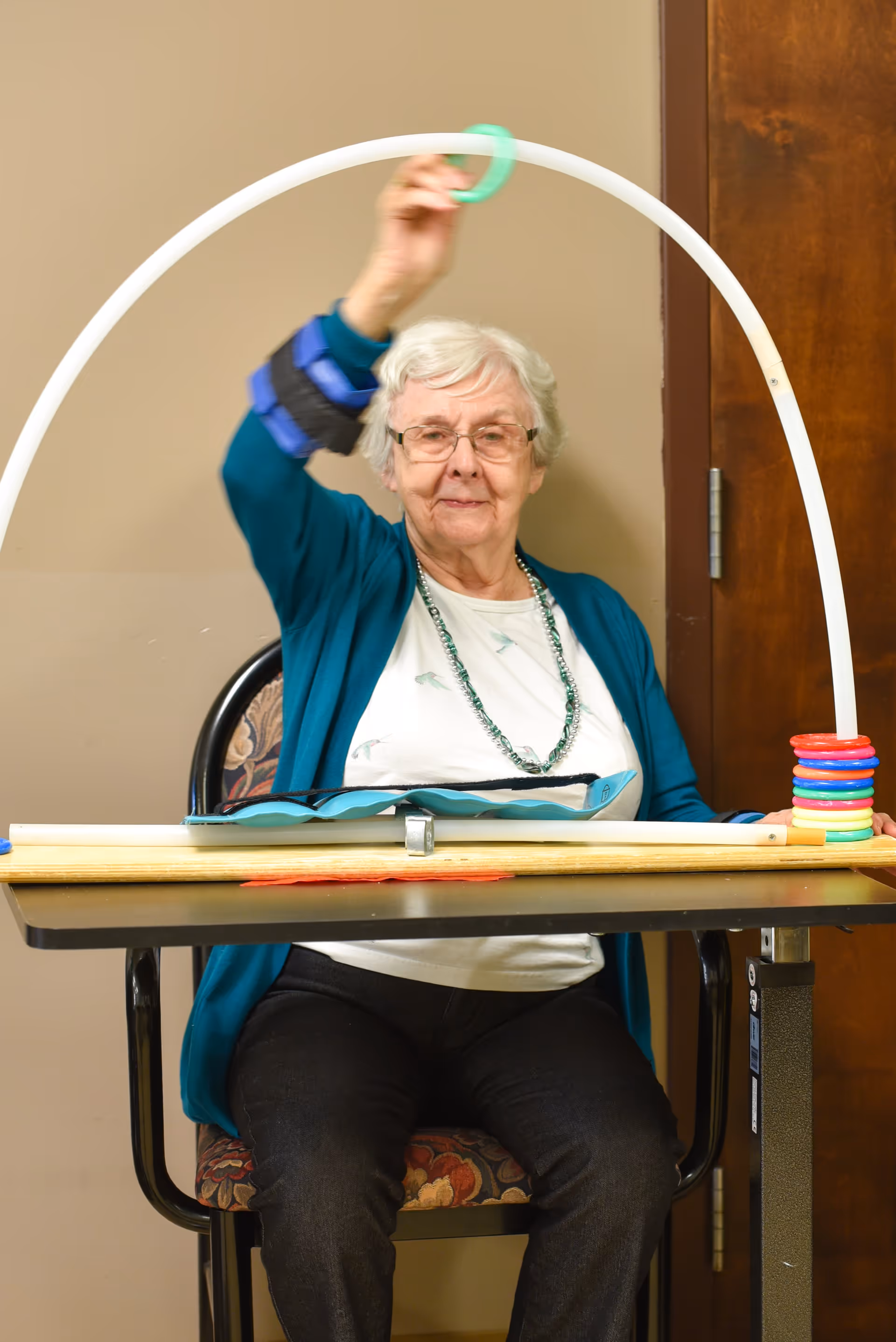 An elderly woman with white hair and glasses is sitting on a floral-patterned chair at a table. She is wearing a teal cardigan over a white shirt and a beaded necklace. She is holding a green ring and appears to be playing a ring toss game with colorful rings stacked on a wooden base with a white curved pole.